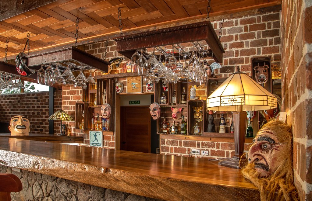 A cozy, rustic-style indoor bar featuring a polished wooden countertop, shelves of spirits, and vintage-style lighting.