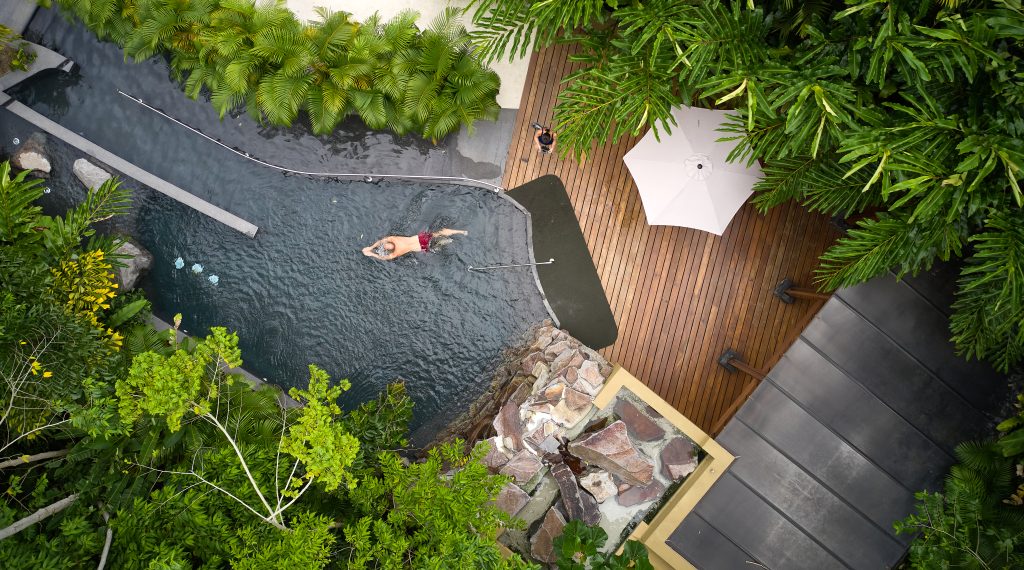 A top-down view of a guest relaxing on a wooden deck next to a private, circular plunge pool surrounded by tropical trees.