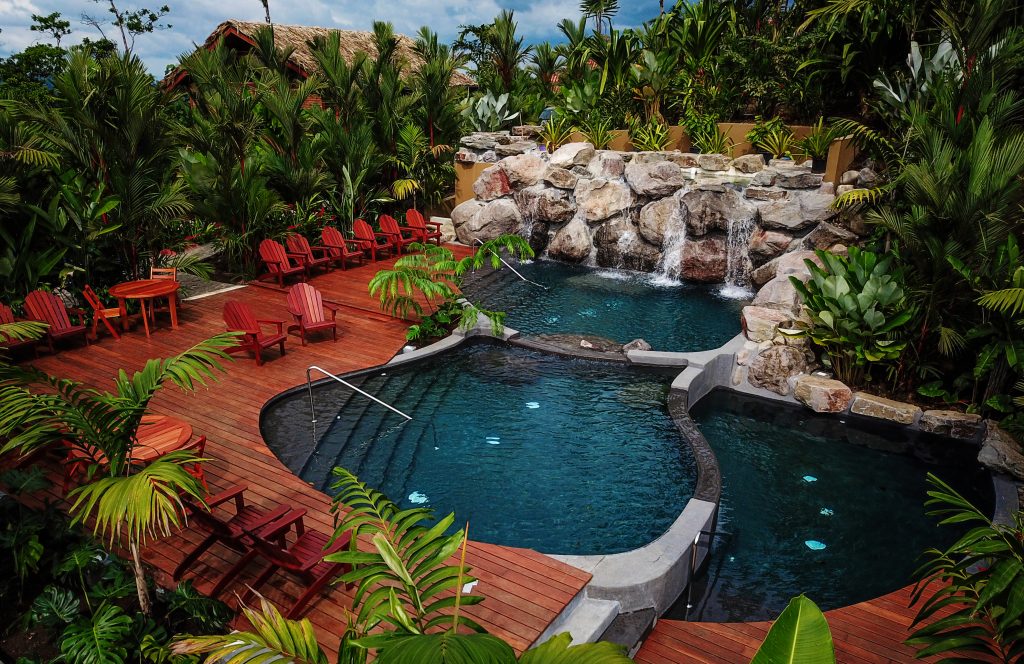 Multi-level outdoor swimming pools featuring rock waterfalls, red lounge chairs, and vibrant tropical landscaping.