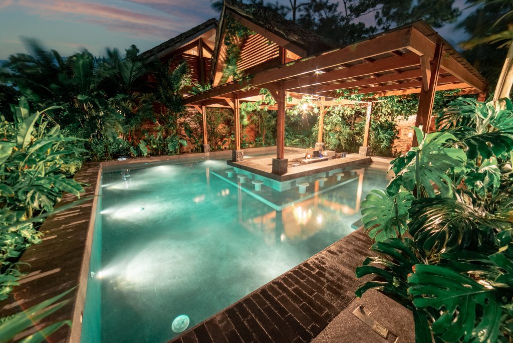 A beautifully lit outdoor infinity pool at night, featuring warm lighting, wooden decking, and surrounding jungle foliage.