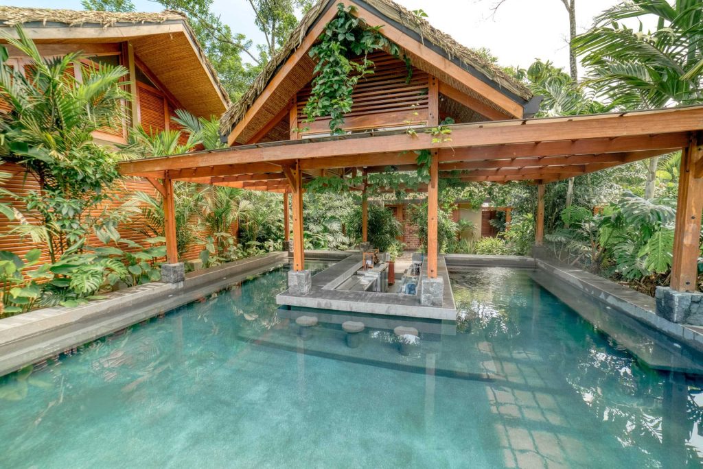 A clear blue swimming pool area sheltered by a large timber-frame pavilion, set against a backdrop of lush tropical greenery.