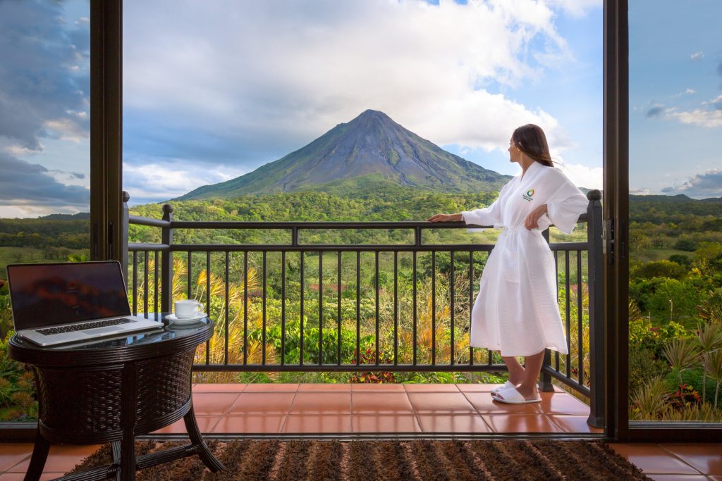 guest in a white robe standing on a private wooden balcony, enjoying a direct and unobstructed view of the Arenal Volcano.