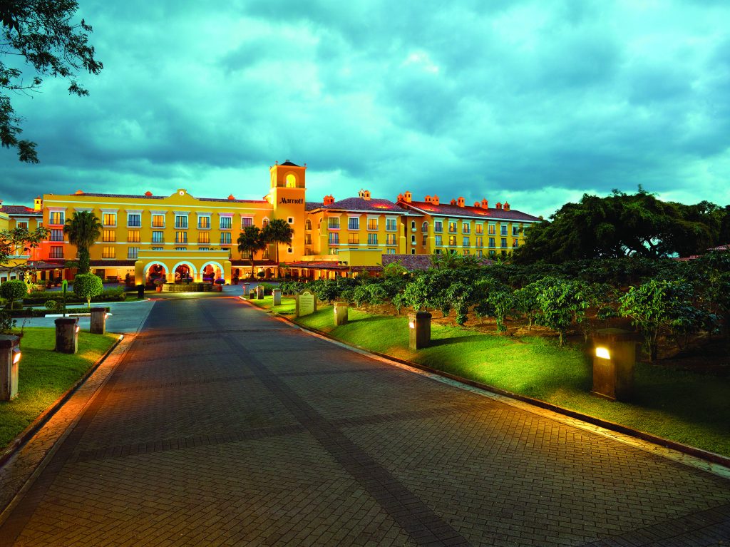 A wide shot of the Marriott Hacienda Belén at dusk, featuring a lit-up colonial-style building and a paved entrance path lined with garden lights.
