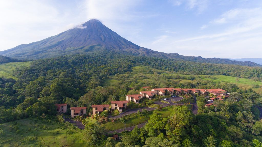 A wide landscape shot showing the majestic, cone-shaped Arenal Volcano rising above the resort&rsquo;s red-roofed villas and green forest.