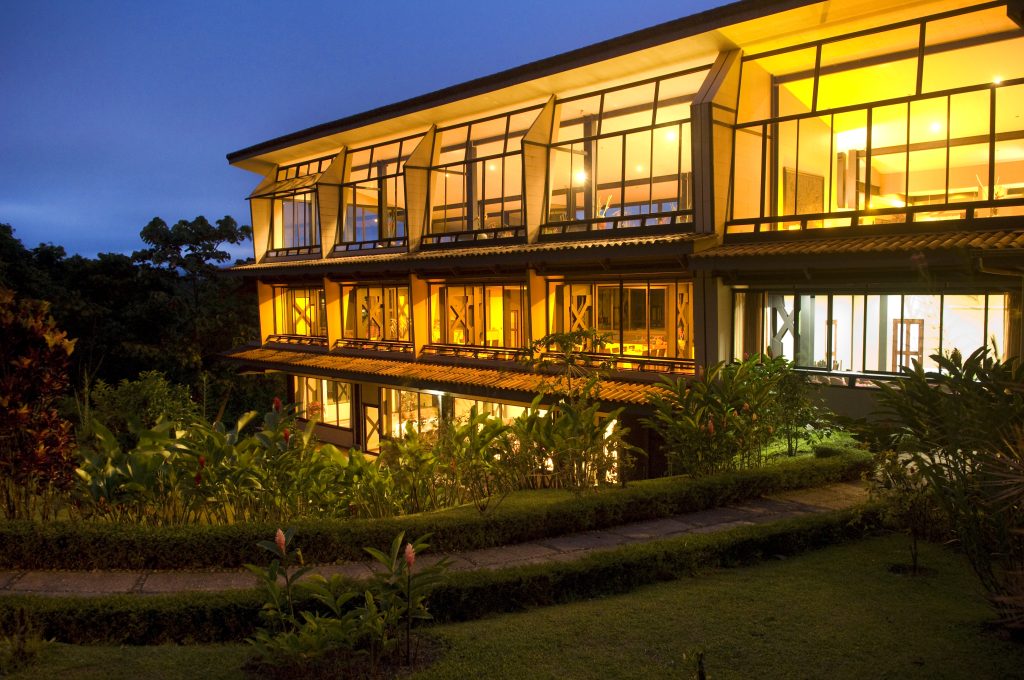 A multi-story hotel wing with large glass windows glowing with warm interior light, surrounded by lush tropical gardens at dusk.