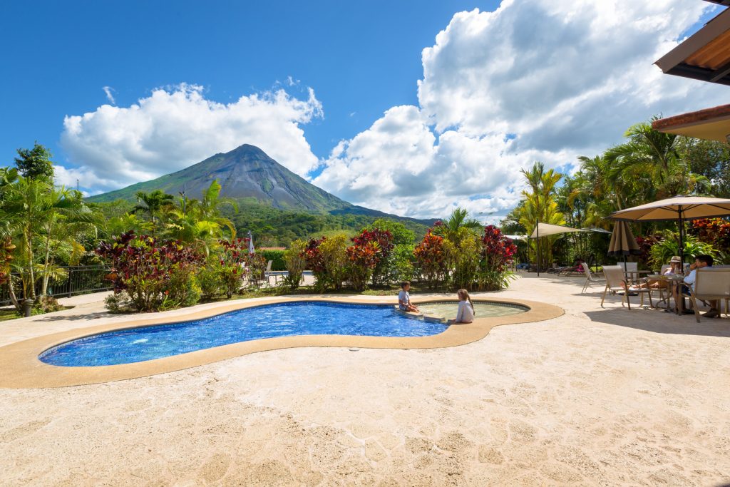 A curved outdoor swimming pool with clear blue water and guests relaxing on the deck, featuring the Arenal Volcano in the background.