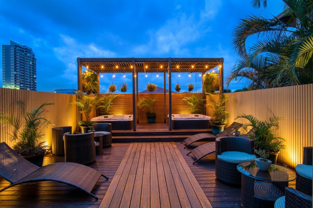 A nighttime view of the rooftop terrace with a wooden deck, comfortable lounge chairs, and an illuminated jacuzzi area.