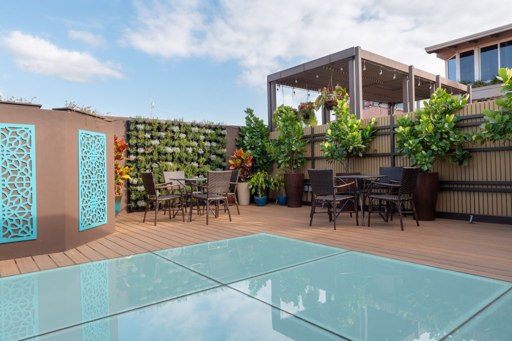 An open-air rooftop space featuring lush greenery, patio seating, and unique glass flooring sections.