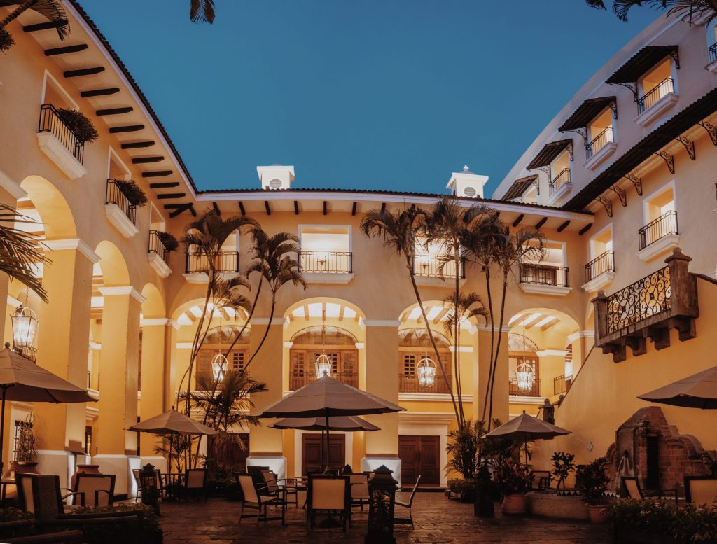 A cozy, illuminated Spanish-style courtyard featuring patio dining, large umbrellas, and indoor-outdoor architecture with palm tree accents.