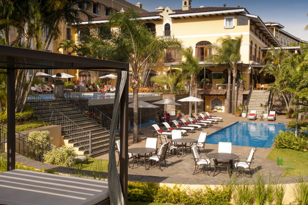 An outdoor swimming pool surrounded by a stone patio with lounge chairs, umbrellas, and dining tables, set against the backdrop of the resort’s architecture.