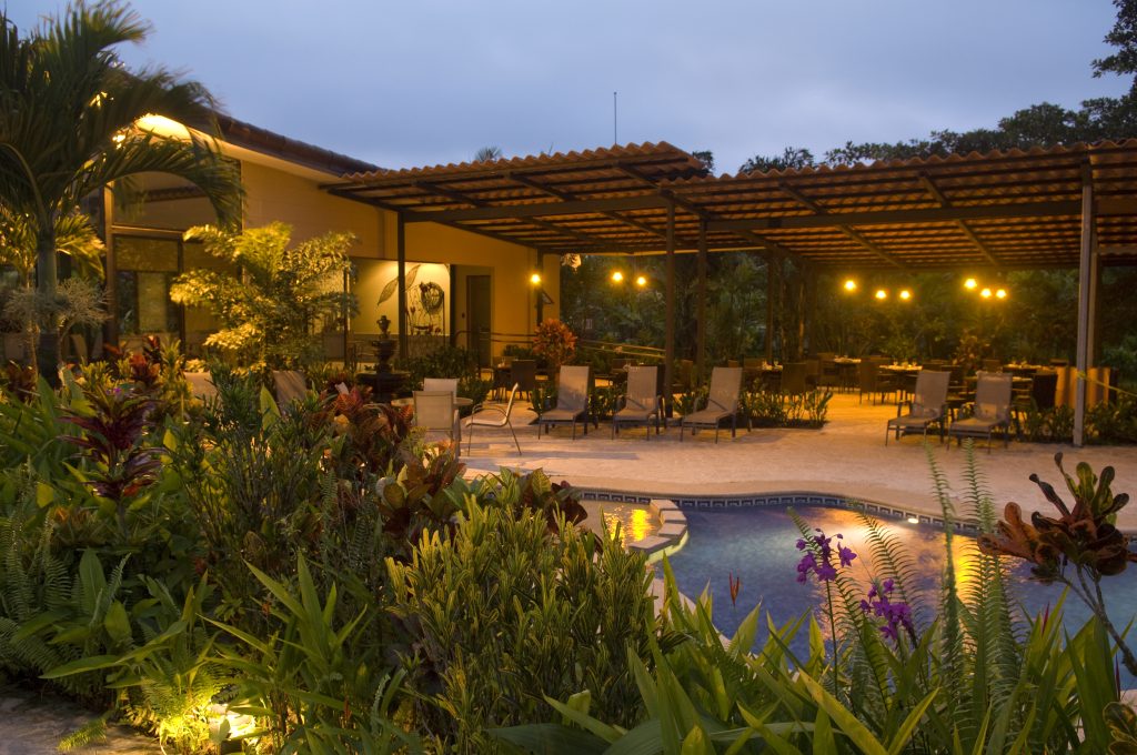 An illuminated outdoor pool area at night, featuring lounge chairs, warm garden lighting, and tropical landscaping.