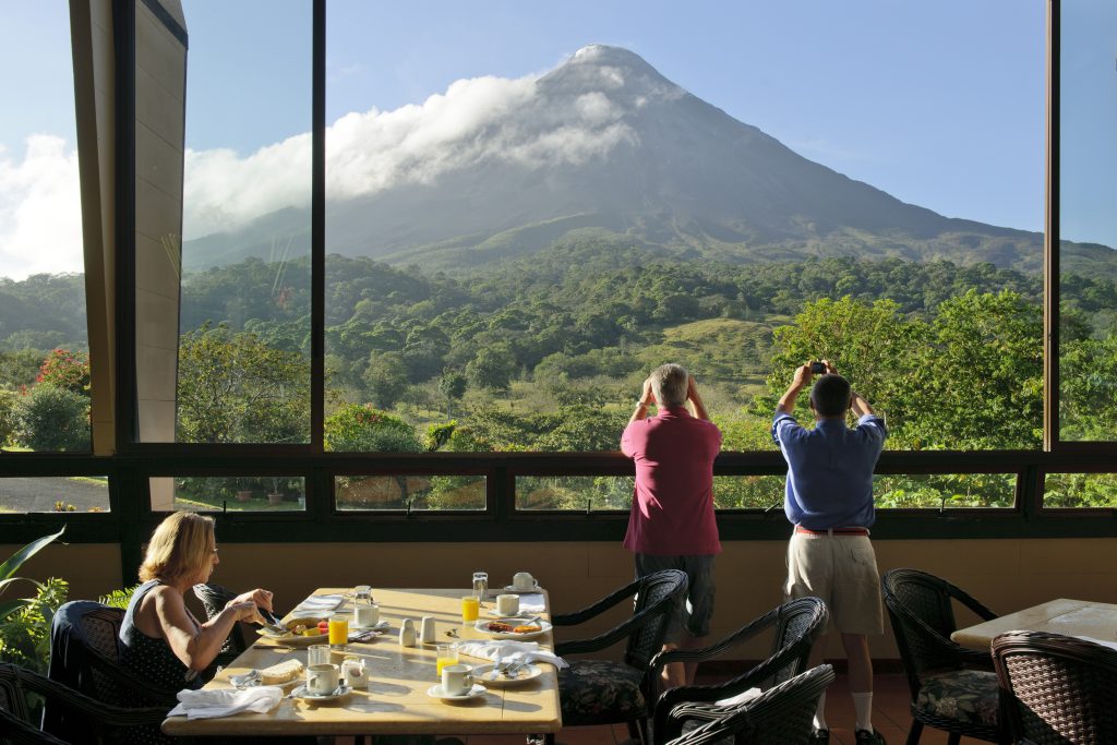 Guests inside a restaurant with floor-to-ceiling glass windows, looking out at the massive Arenal Volcano while dining.