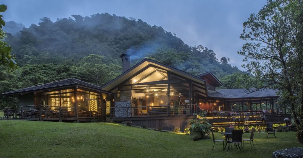 Exterior view of the main lodge at El Silencio at dusk, featuring warm lighting and a foggy mountain backdrop.